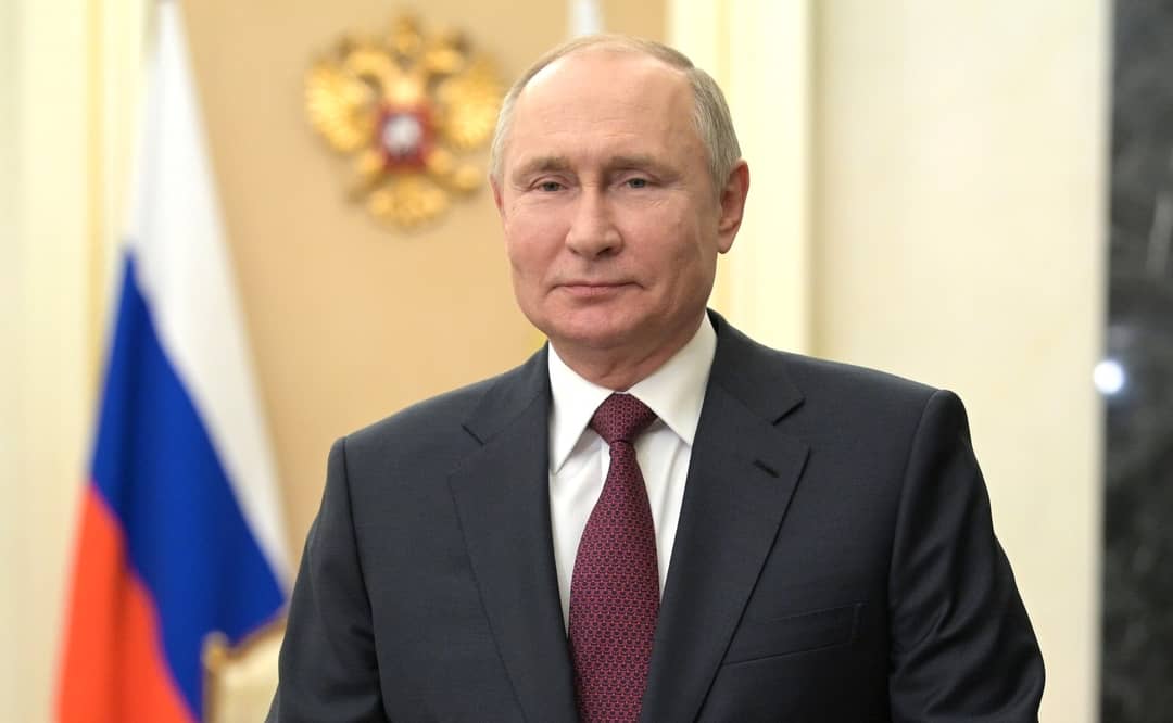 Vladimir Putin stands in front of a Russian flag.