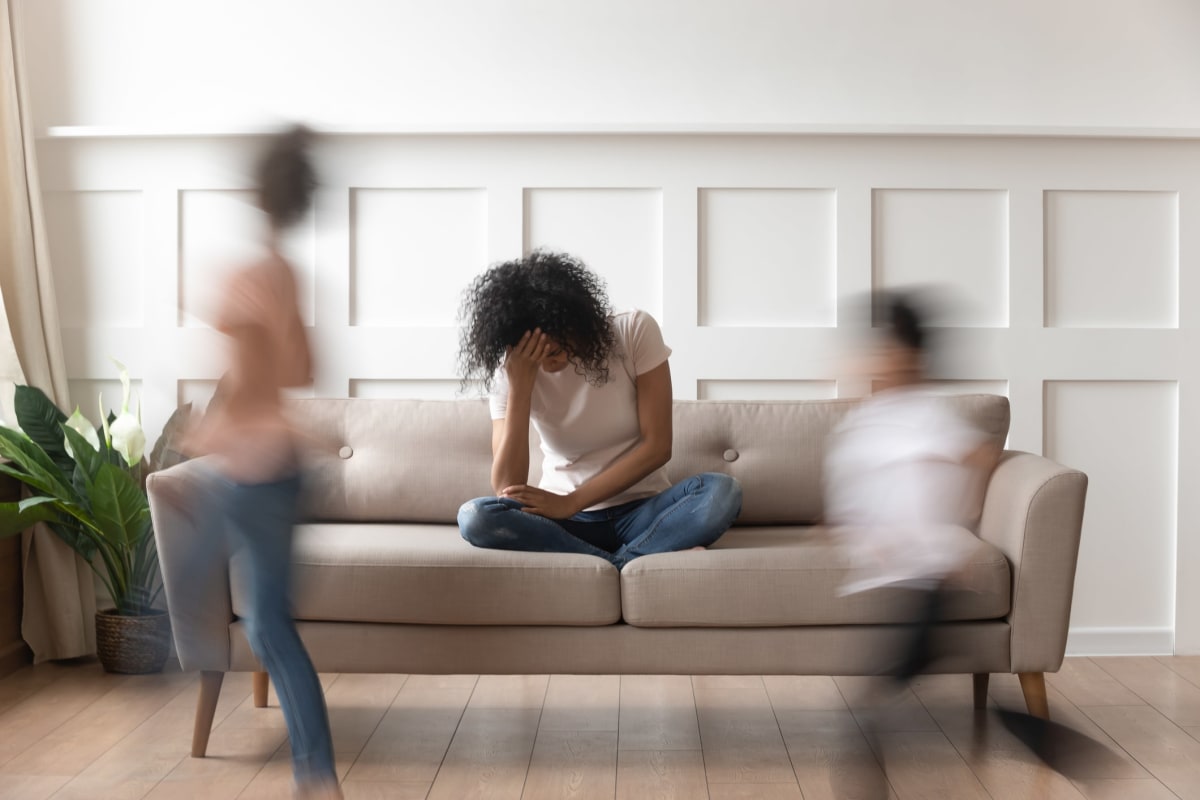 Stressed mother on a couch with head in had as blurred children run around