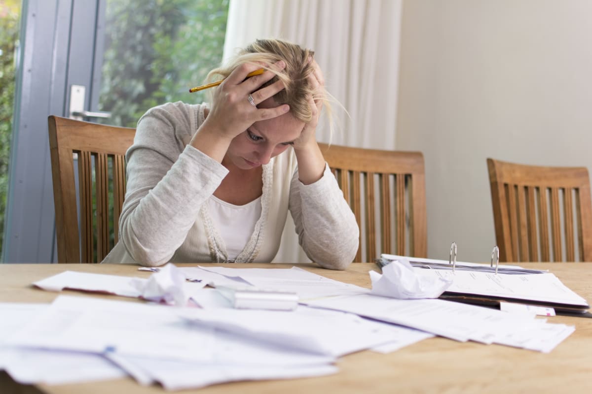 A woman sits at a table with her head in her hands, surrounded by bills on a table.