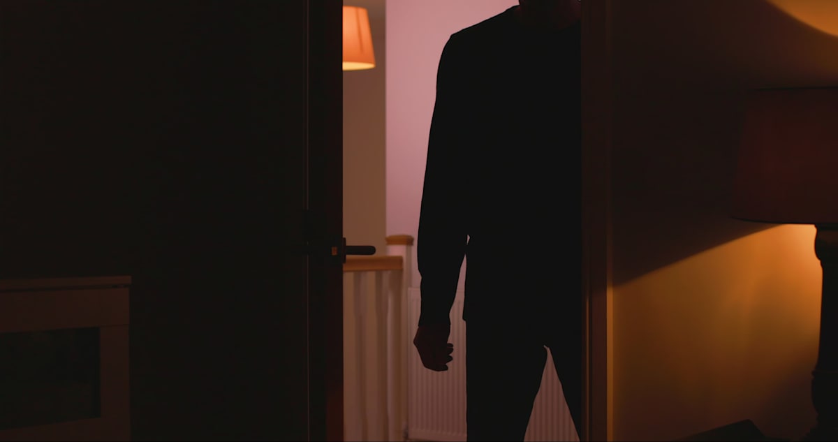 Silhouette of a man standing in an open doorway leading to a dark room with a side lamp