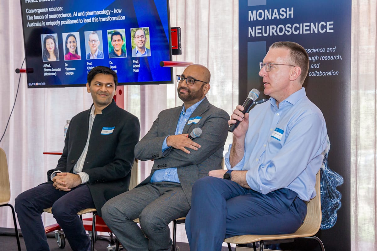 A seated Dr Deval Mehta, Professor Adeel Razi and Professor Chris Langmead, who holds a microphone and is speaking, at the neuroscience showcase
