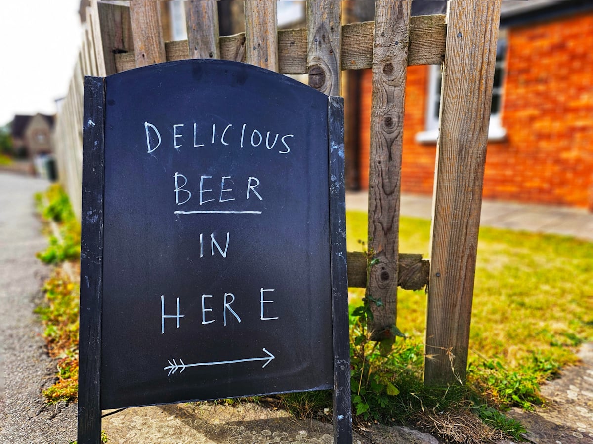 A-frame sign on a sidewalkl with the words delicious beer here written on it.