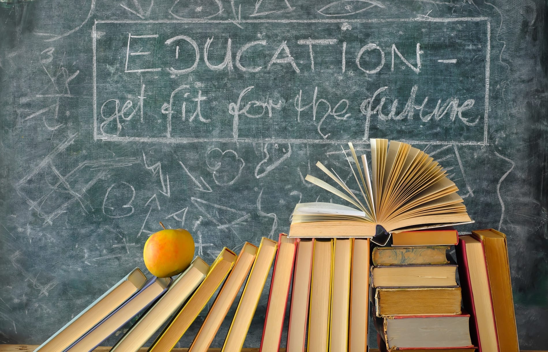 Books and and apple on a school desk with a blackboard in the background with writing on it.