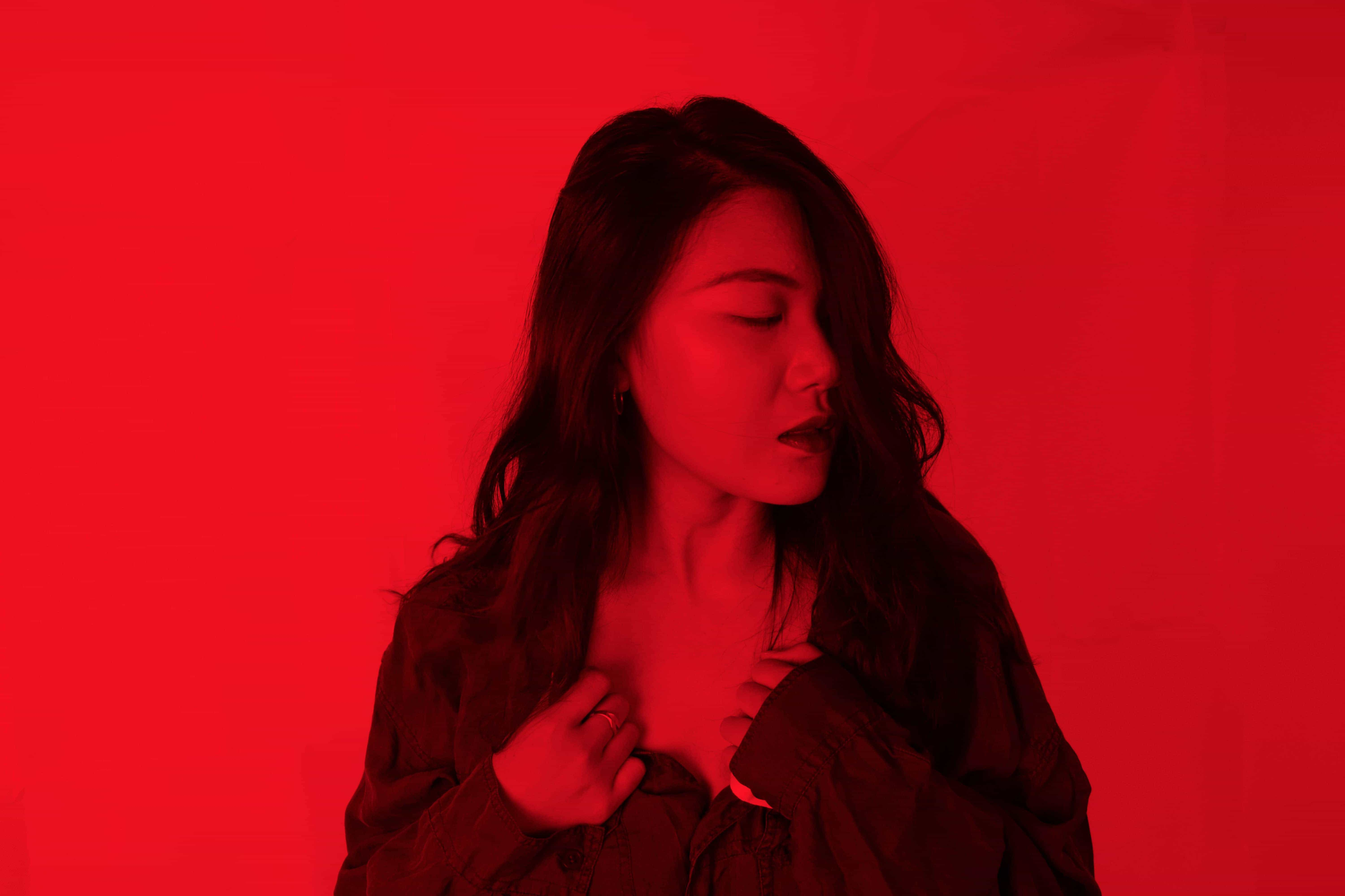 A woman clutching at the neck line of her shirt with eyes closed in contemplation with the photo having a red overlay.