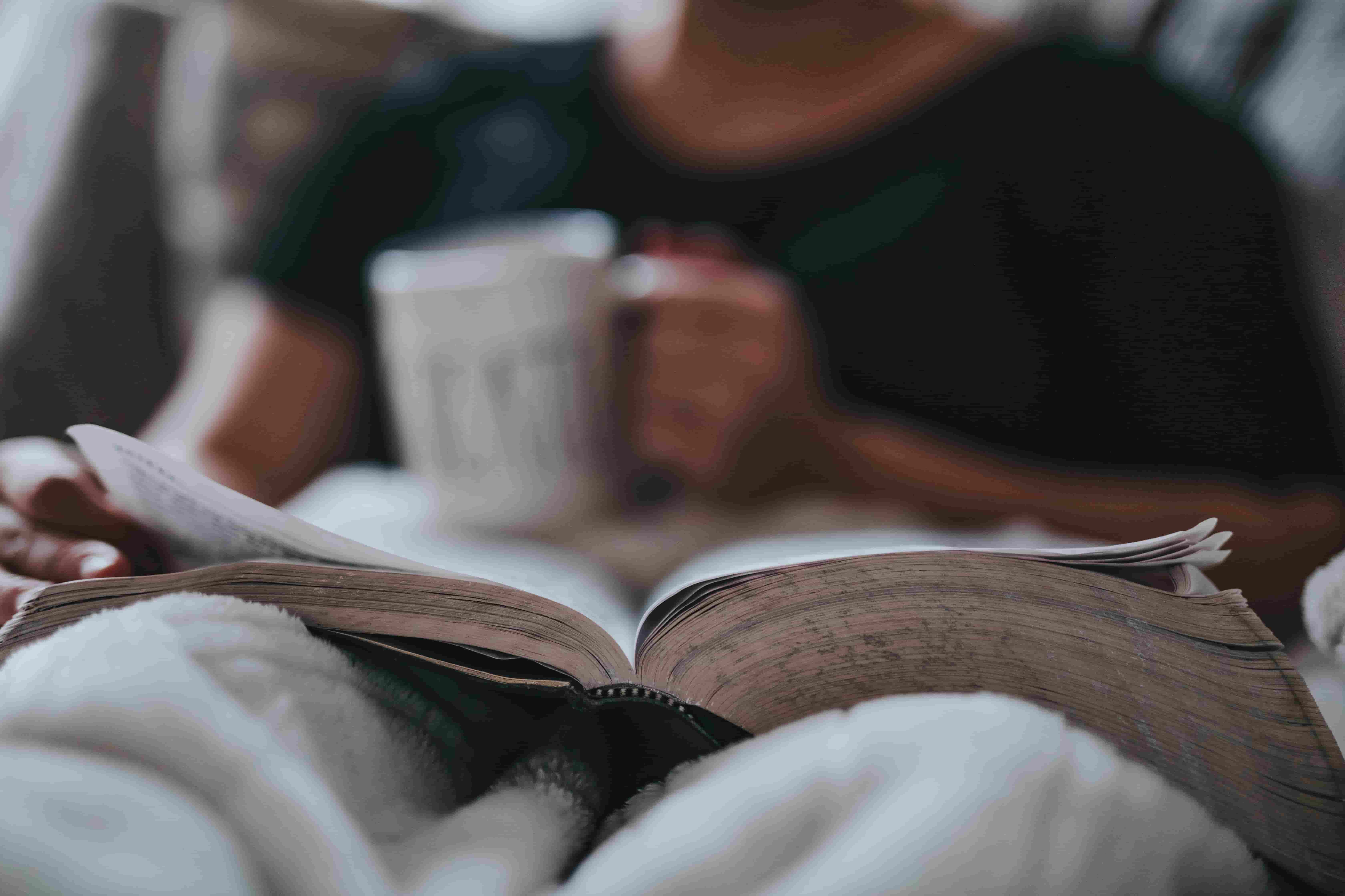A person sitting on a bed or couch and holding a coffee mug in one hand and reading a book an dturning a page with the other hand.