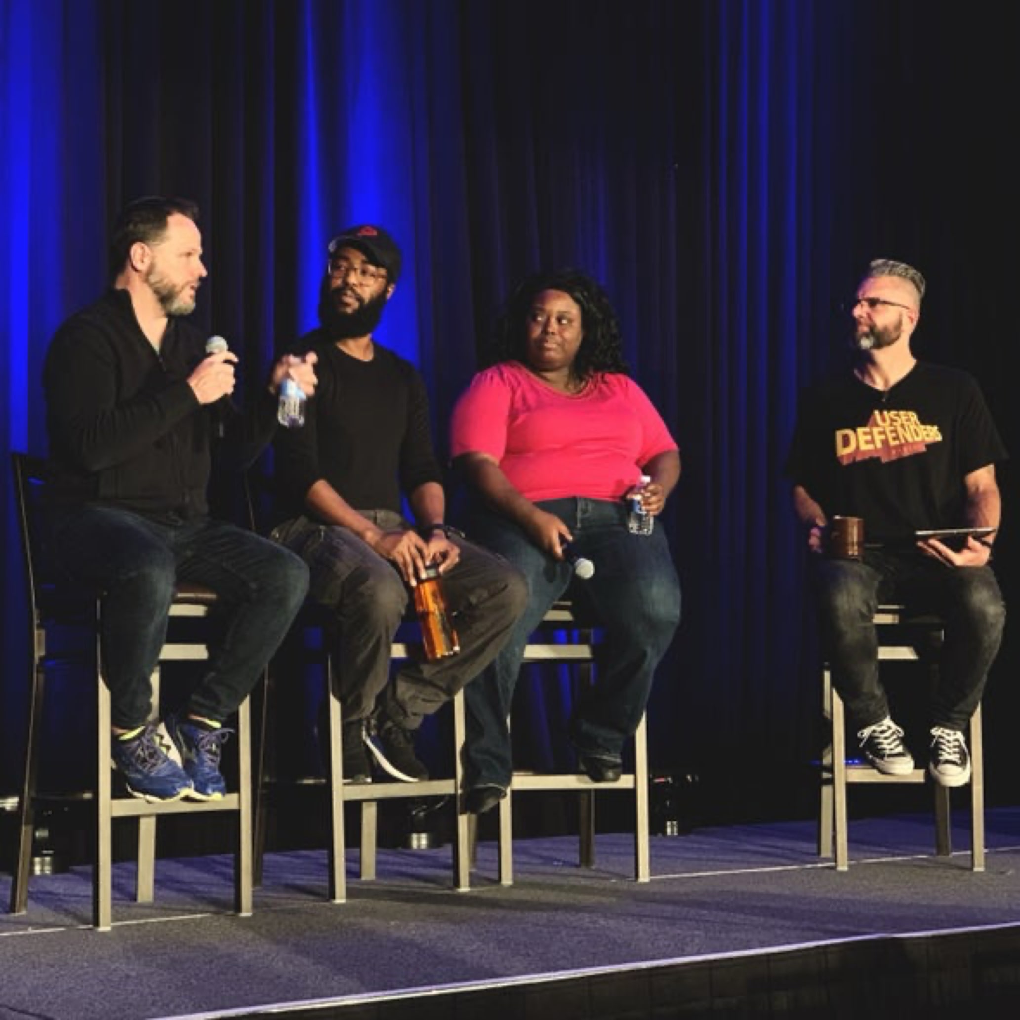 User Defenders Podcast live on stage at An Event Apart Denver. From Left to right, Derek Featherstone, Farai Madzima, Mina Markham and Jason Ogle sitting on stage on stools with a blue curtain backdrop. Photo courtesy of Jeffrey Zeldman