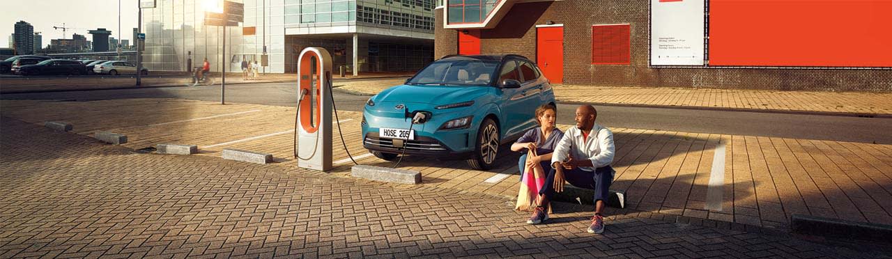 יונדאי קונה חשמלית בצבע כחול מחוברת לעמדת טעינה ולידה יושבבים גבר ואישה על מדרכה - מבט על חזית הרכב
