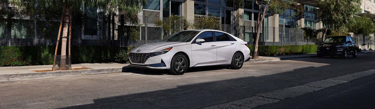 יונדאי אלנטרה היברידית בצבע לבן חונה בצד בכביש תחת בניין ועצים