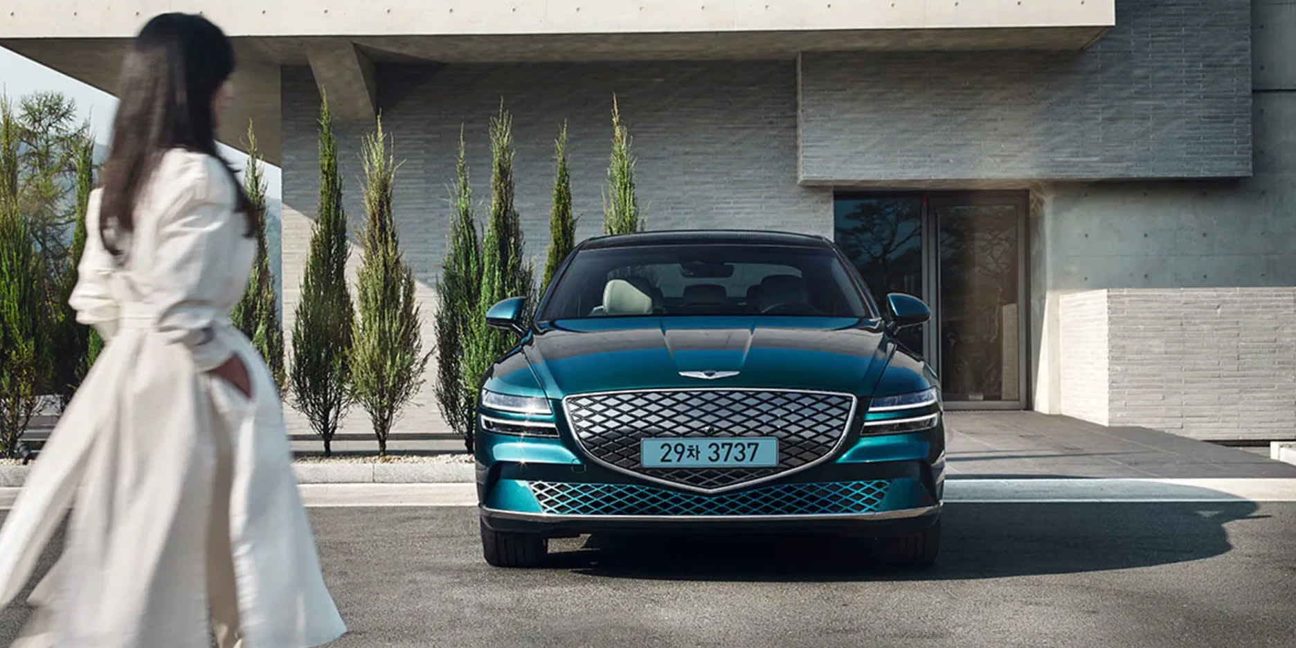 תמונת אווירה לצורת מאמר השוואת רכבים חשמליים - רכב מדגם Electrified G80 בצבע כחול מטאלי חונה ואישה מתקרבת אליו