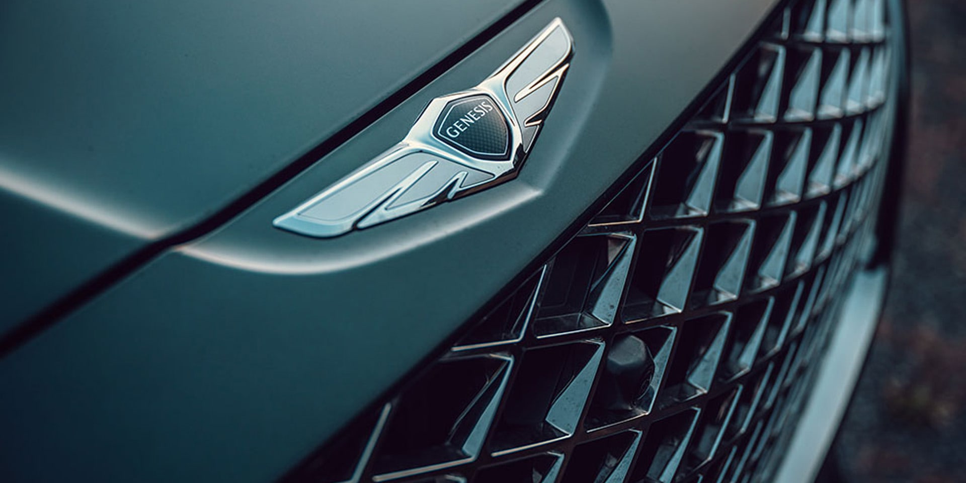 תקריב על סמל ג'נסיס על מכסה מנוע רכב יוקרה, לצד גריל קדמי בעיצוב גיאומטרי