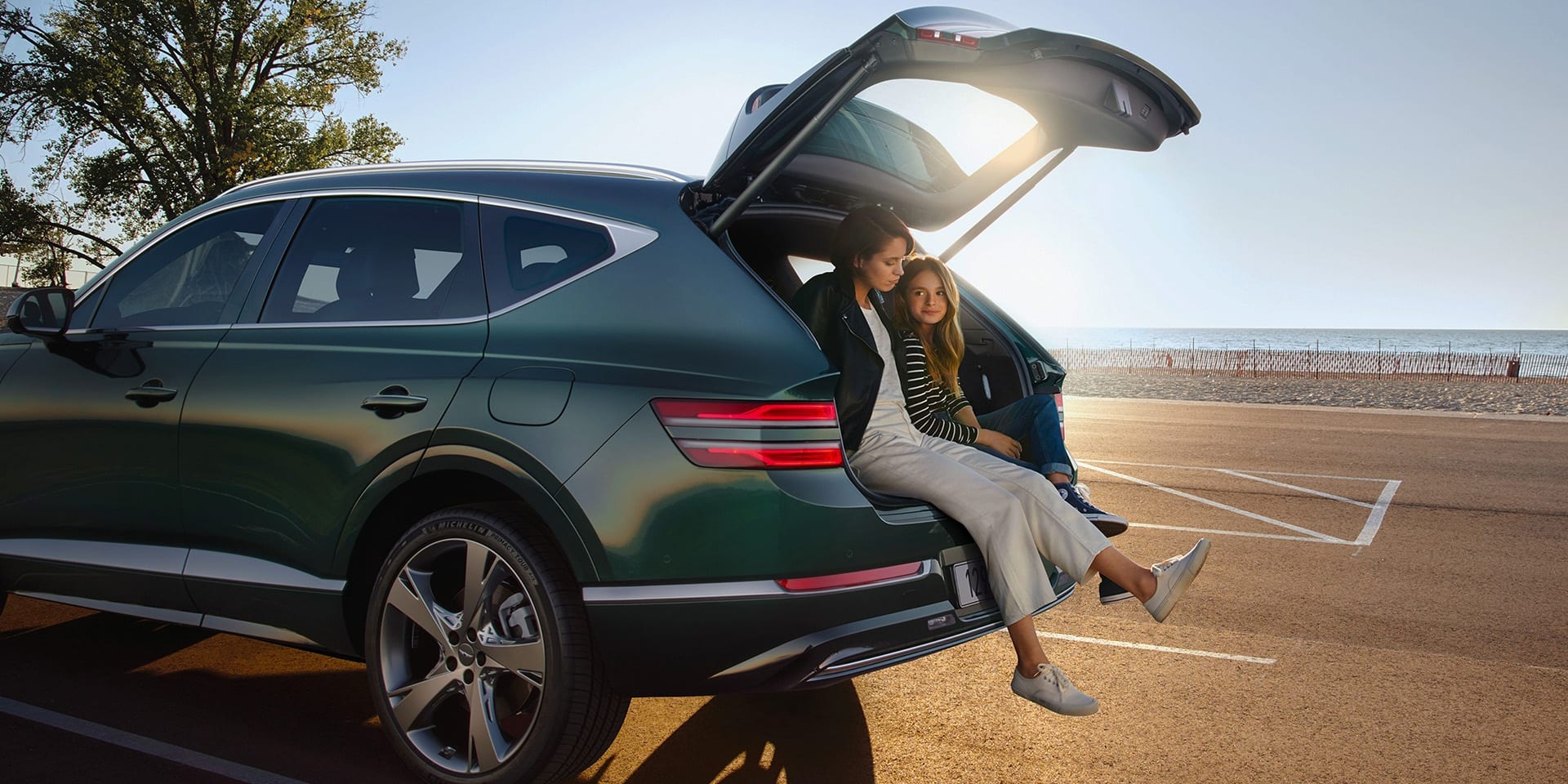 אם ובת יושבות על תא המטען הפתוח של רכב יוקרה ג'נסיס ירוק, בחנייה מול חוף ים
