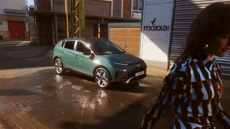 יונדאי באיון חונה על מדרכה בסביבה עירונית ואישה מתרחקת מהרכב - מבט על חזית וצד הרכב