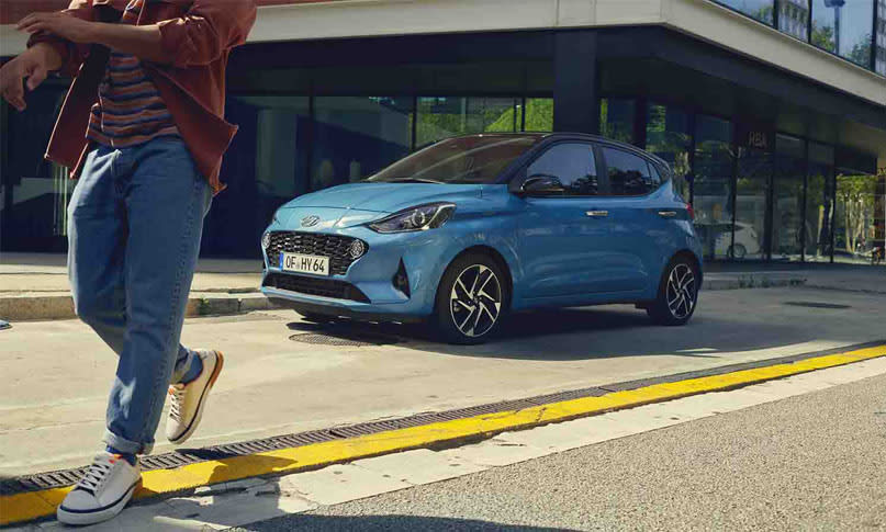 יונדאי i10 בצבע כחול חונה בצידי הכביש וגבר מתרחת ממנה - מבט על חזית וצד הרכב