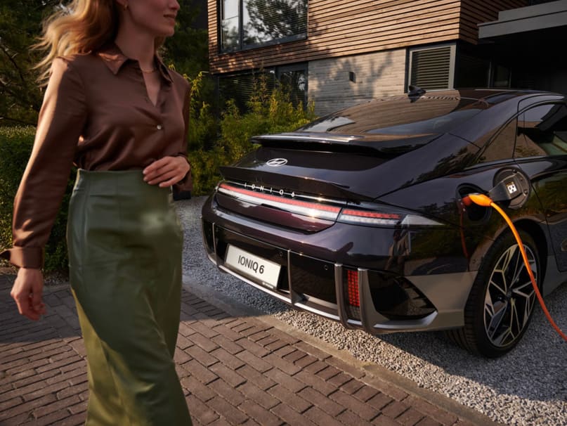 רכב חשמלי מחובר לטעינה, אישה עוברת ליד הרכב