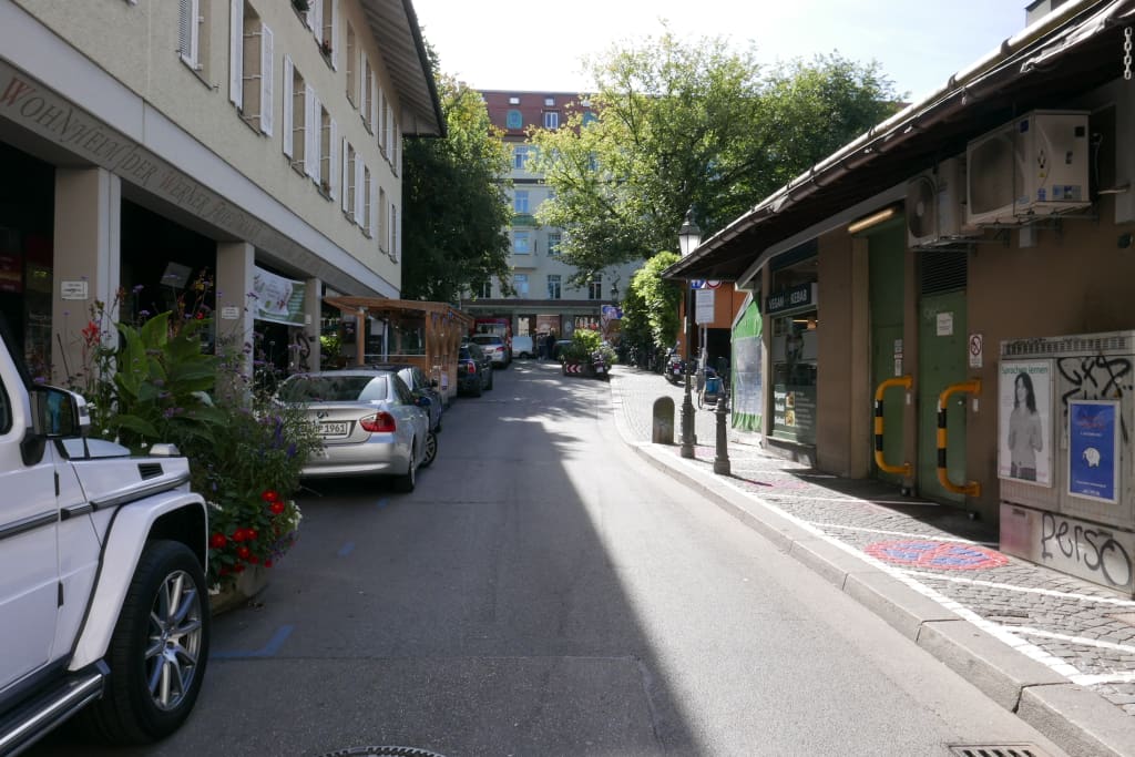 View up a road in Munich city entre with parked cars on one side, and bollards containing plants and flowers placed intermittently down the road, with signals for cars to slow down. 