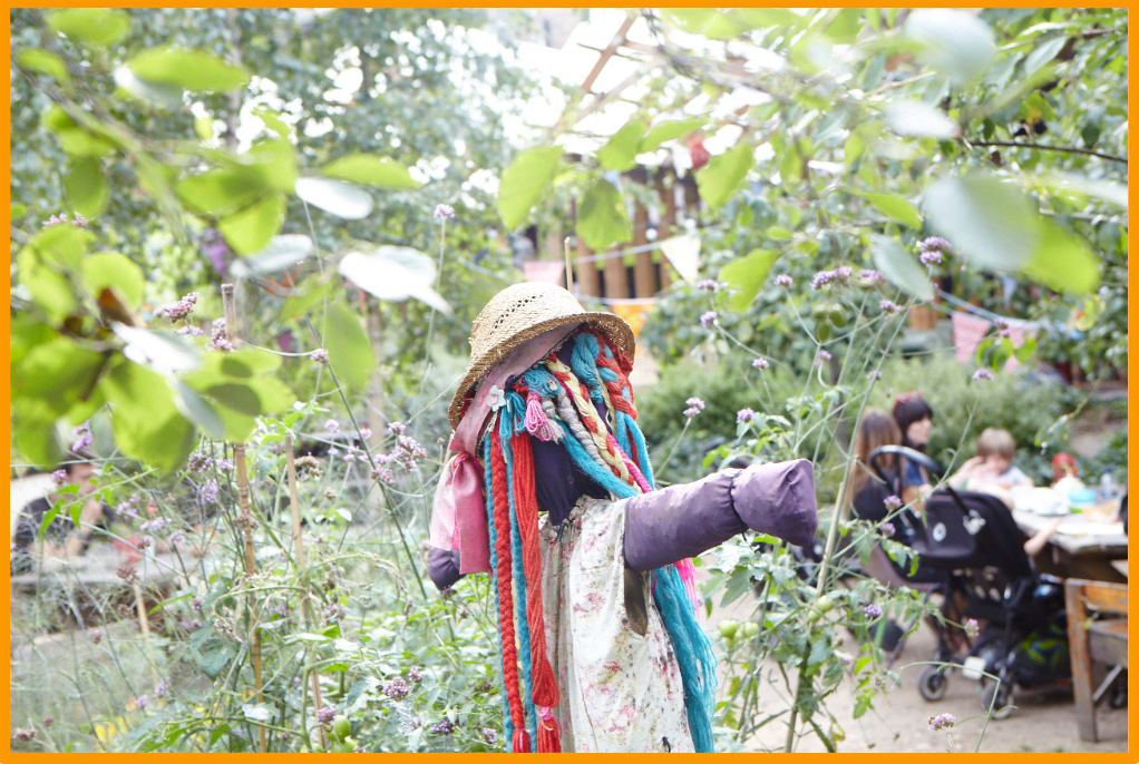 up-close shot of a scarecrow in the curve garden. Two ladies with baby sitting on a table in the distance 