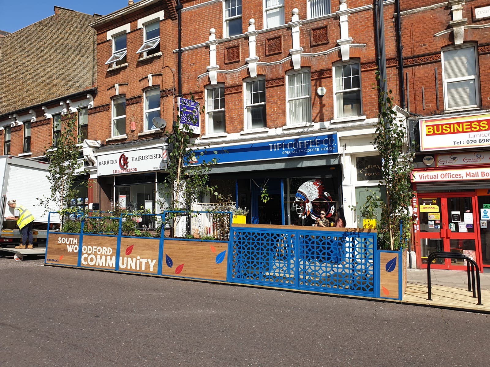 Photo of the entire Mobility Hub wooden structure, including trees and plants, cycle parking stands, blue metal fencing and detailing, South Woodford Community written in big white letters.