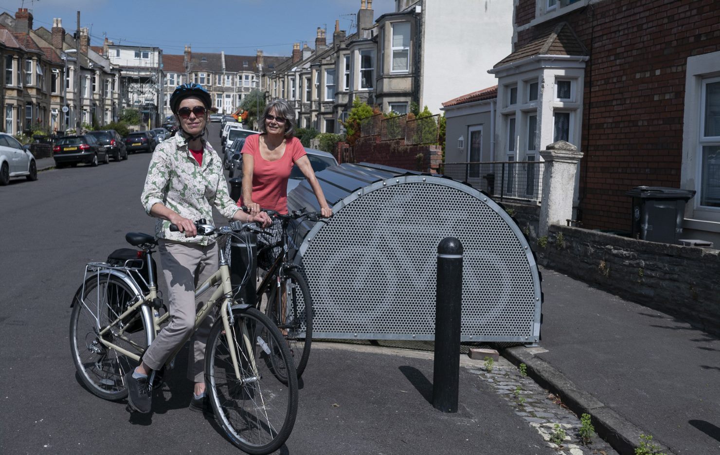 A man and a woman on their bikes next to a cycle hangar on the side of the road next to the pavement. 