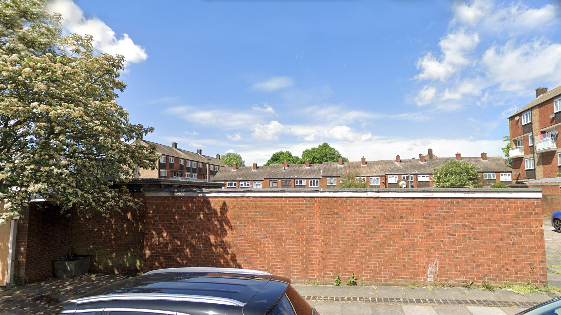 Photo of the site as it is now, with the back of a red brick garage in the foreground, a tree to the left, a block of older flats to the right and terraced houses behind.