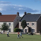 Two houses on a village green with two people walking on the green and someone sat on it.
