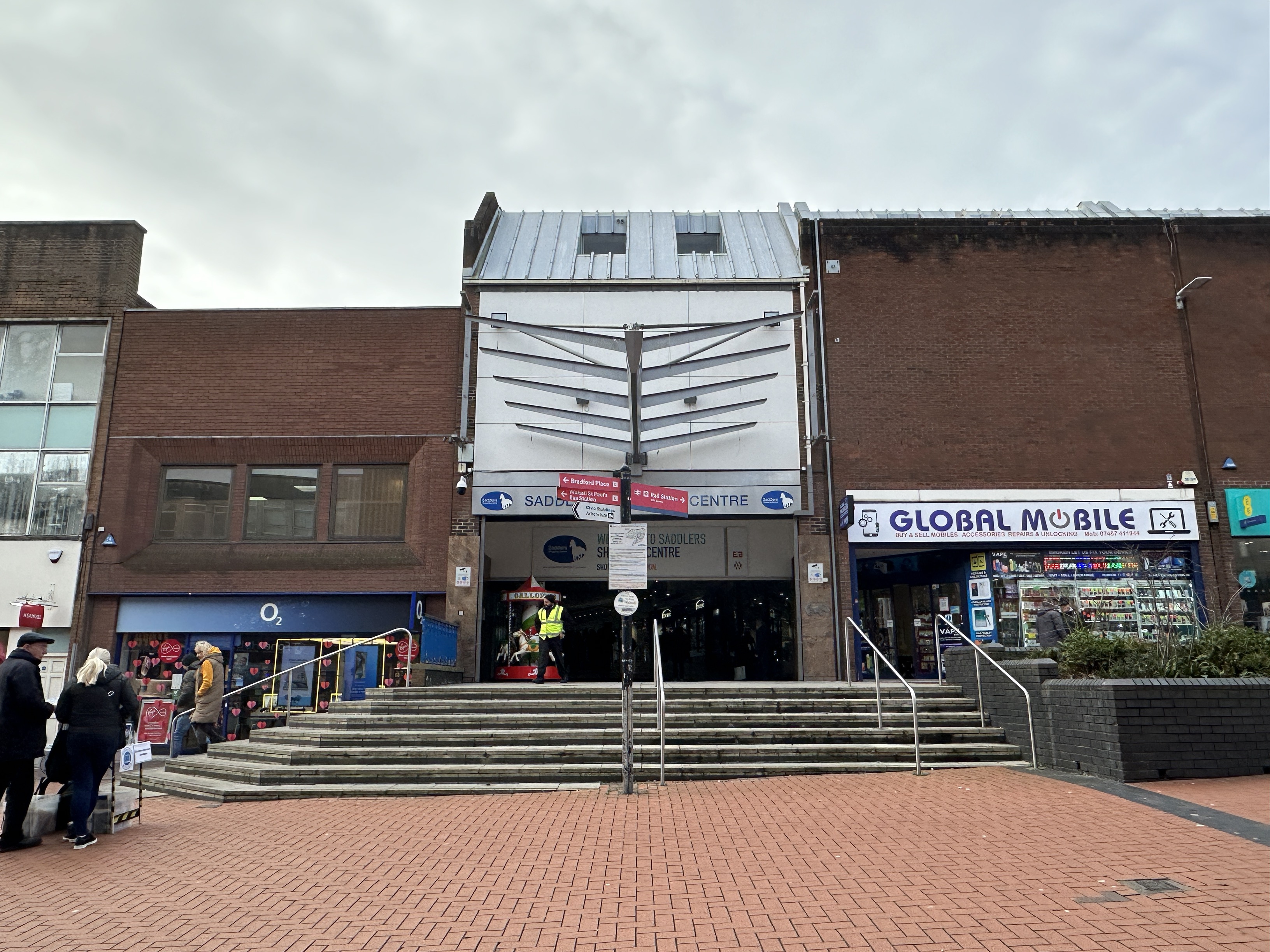 The front of a shopping centre. The ground is red brick and there are grey steps in front of the building. The entrance is also red brick with a pale grey section in the middle above the doors. The sign says 'Saddlers Centre' in blue. There are a few people walking around outside and to the right is a grey brick planter with greenery in.