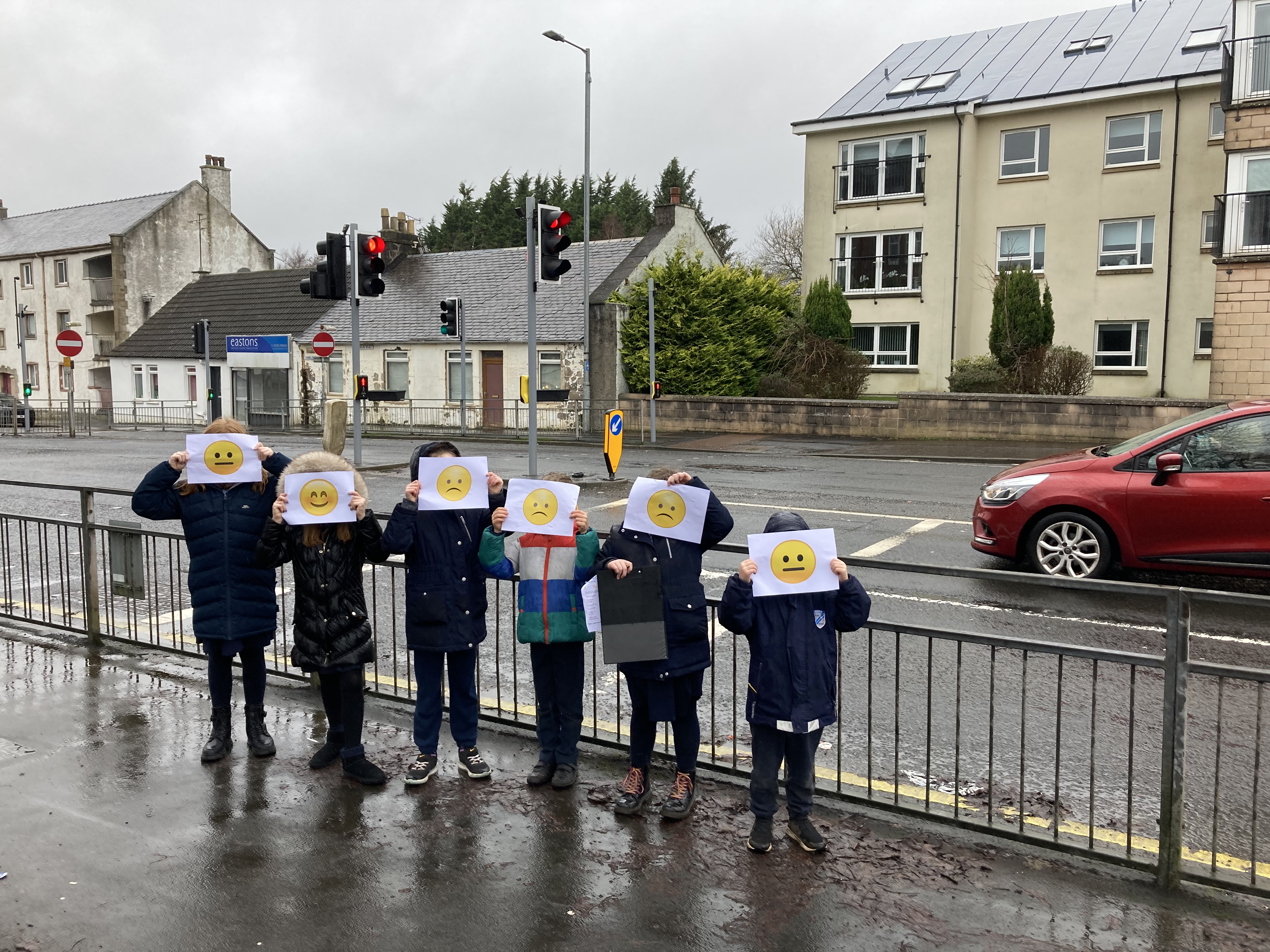 An image of six school children standing in a line on the pavement next to a Main Road holding bits of paper with neutral and sad faces over their faces