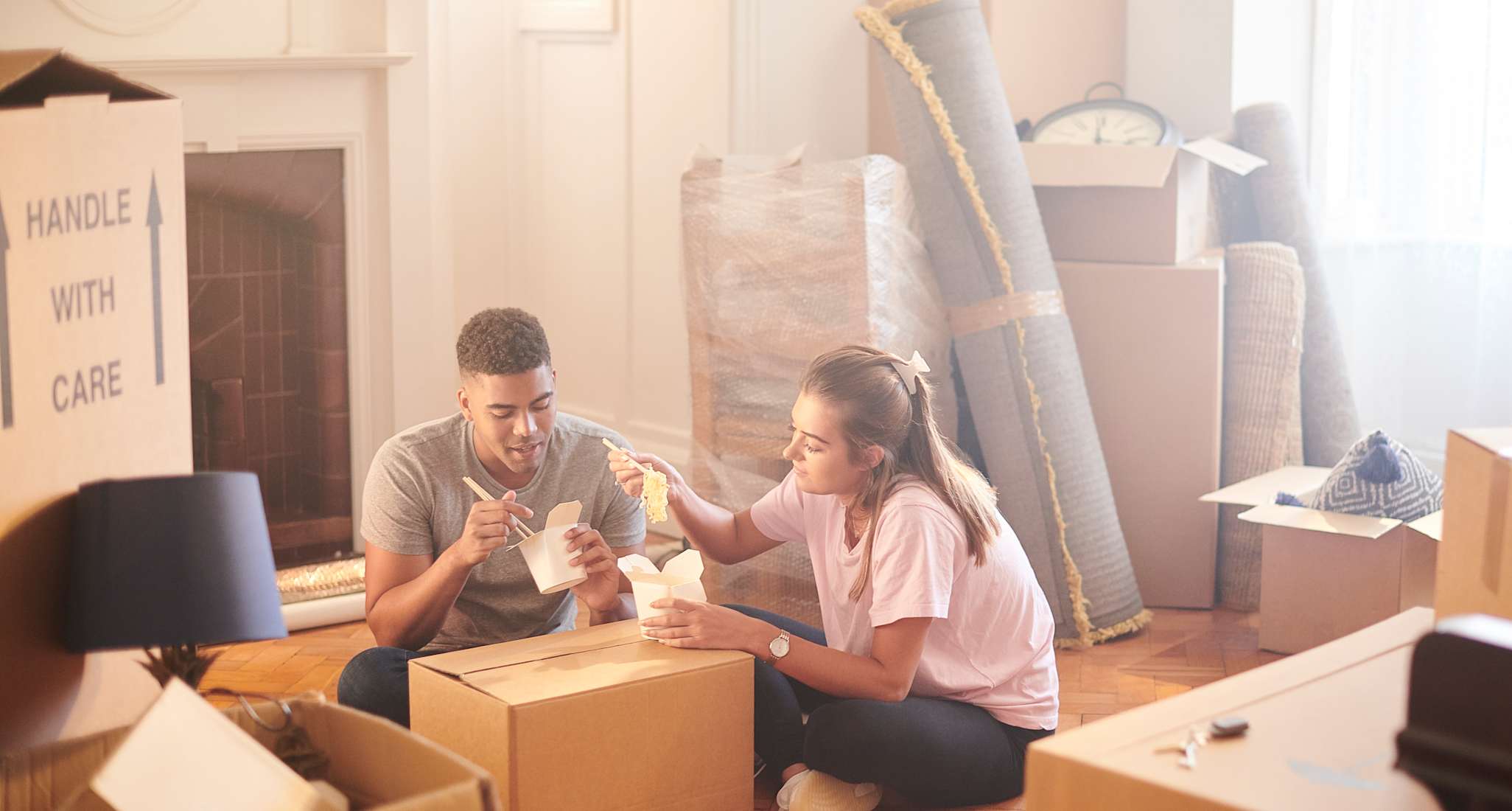 A couple eats take-away noodles surrounded by moving boxes in their new apartment.