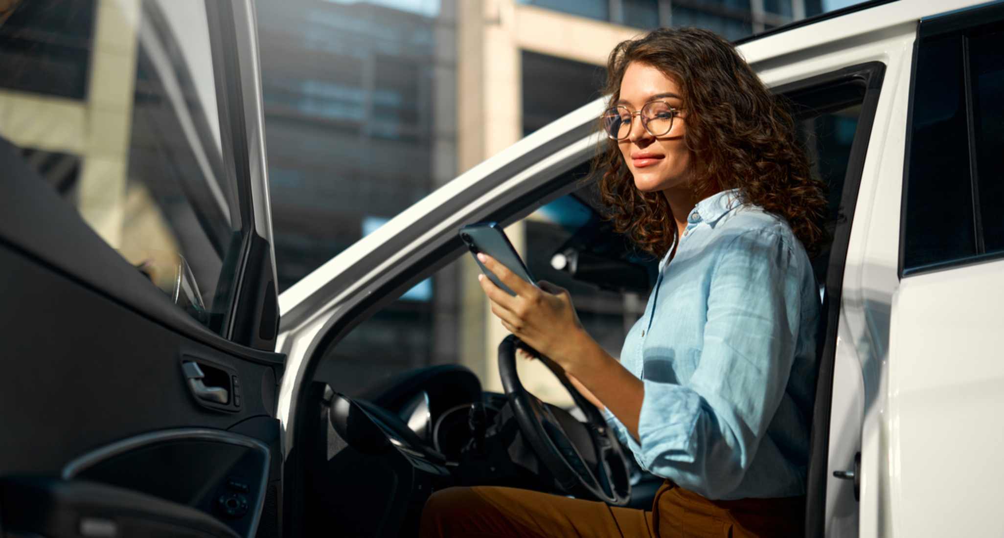 Junge Frau sitzt in einem weissen Auto mit offener Tür und schaut lächelnd auf ihr Smartphone
