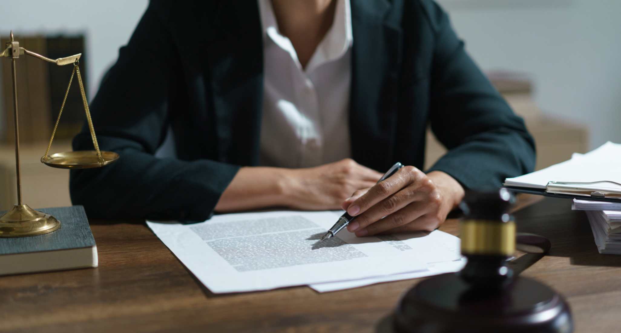 Une avocate en blazer noir signe un document juridique à son bureau avec une balance et un marteau de juge.