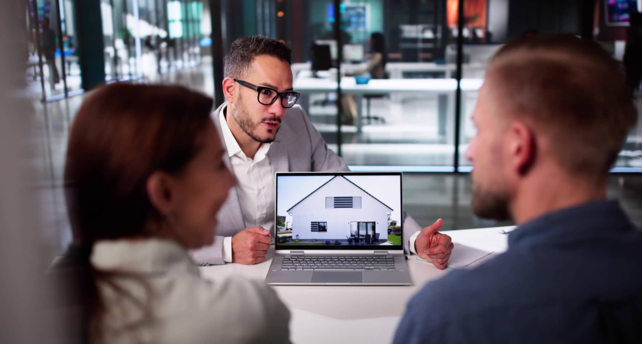 Professioneller Immobilienmakler präsentiert einem glücklichen Paar Immobilienoptionen mithilfe eines Laptops in einer modernen Büroumgebung.