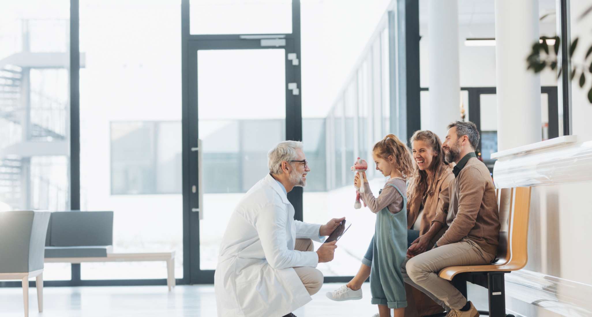Eine Familie im Wartezimmer eines Krankenhauses. Ein Arzt hockt vor der stehenden jungen Tochter.