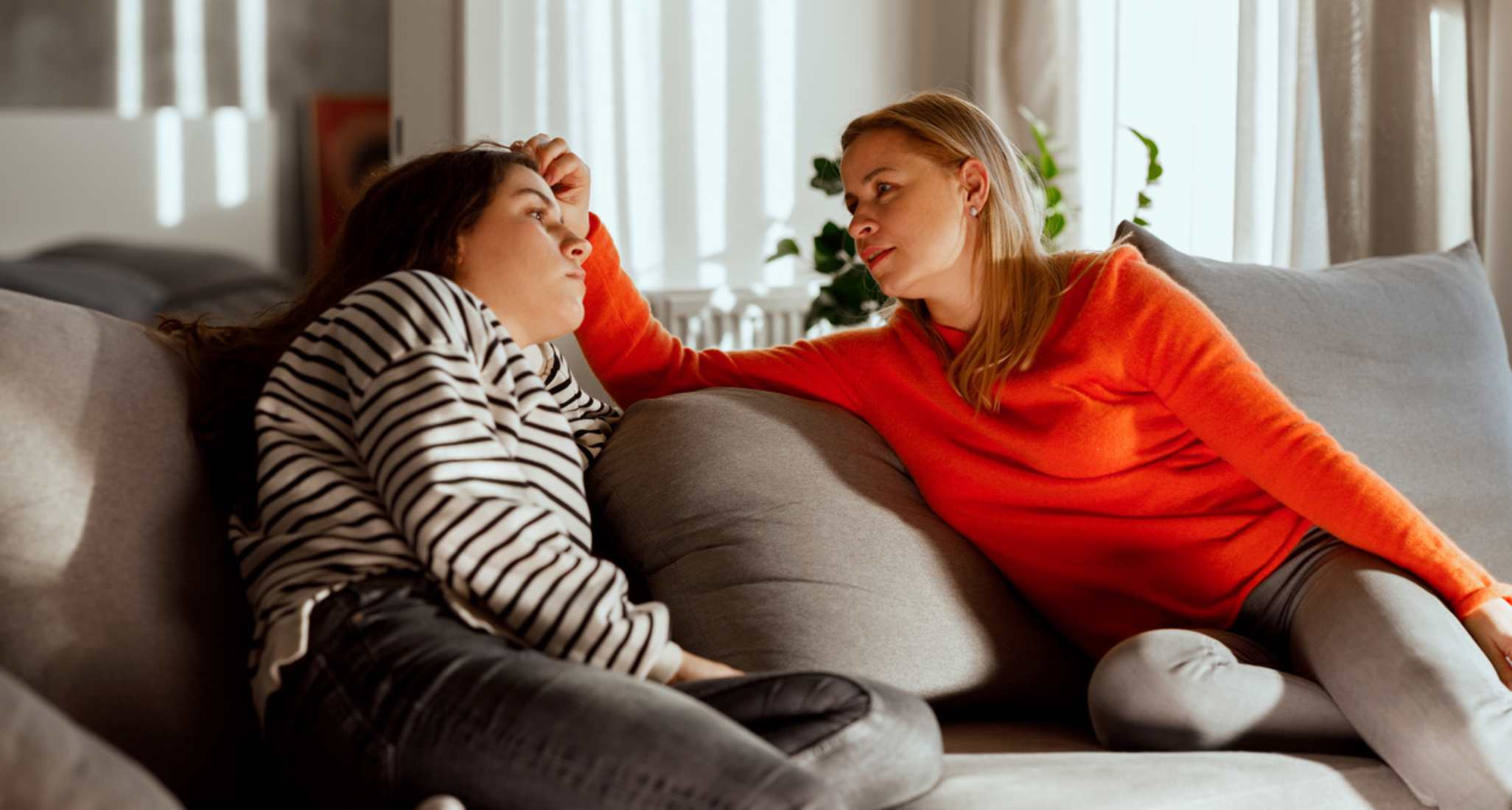 A mother and her teenage daughter sit on a sofa. The mother comforts her worried daughter.
