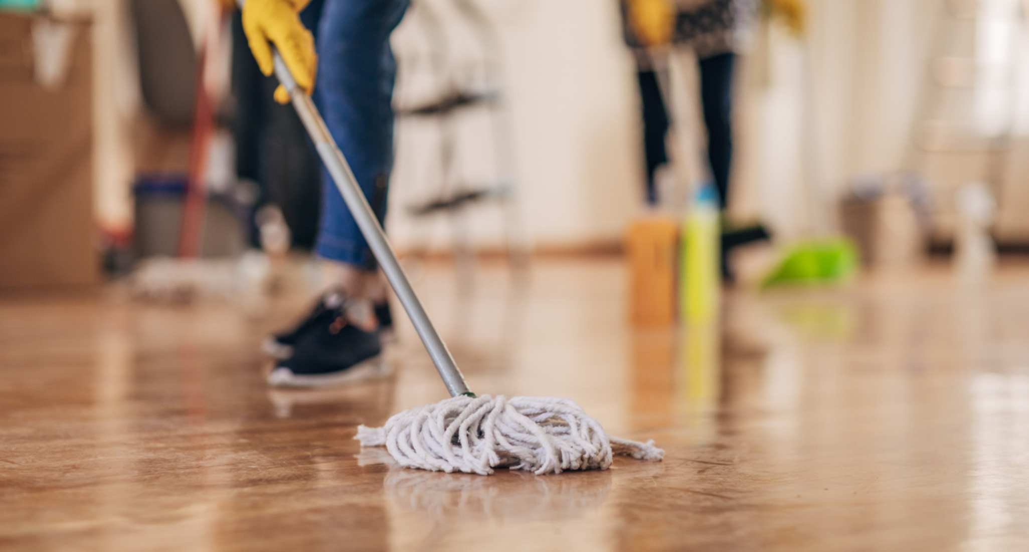 Eine Person mit Mop putzt den Boden. Im Hintergrund ist unscharf eine zweite putzende Person zu sehen.