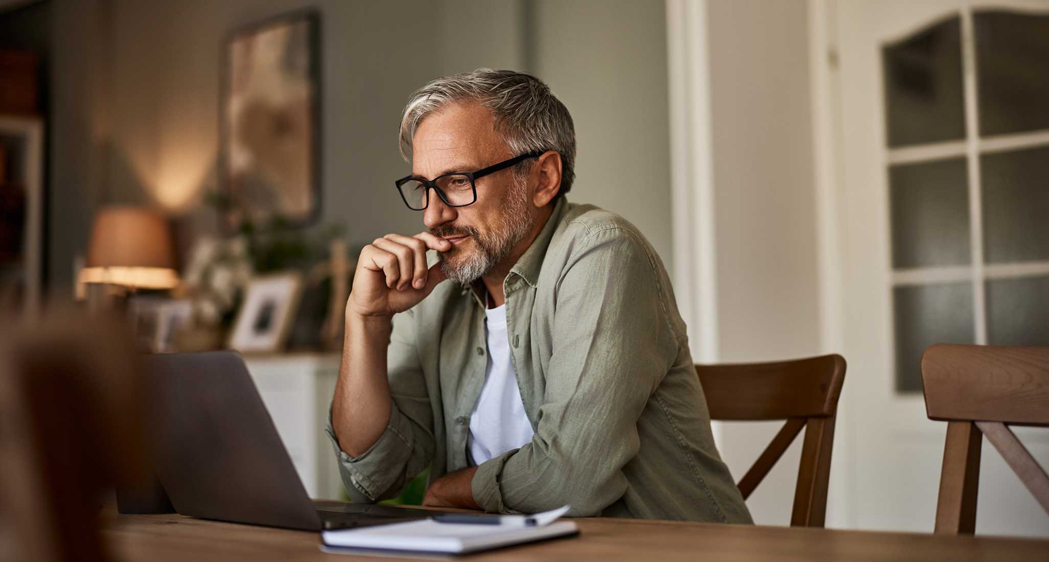 Mann mit Brille sitzt am Esstisch und schaut nachdenklich auf seinen Laptop.