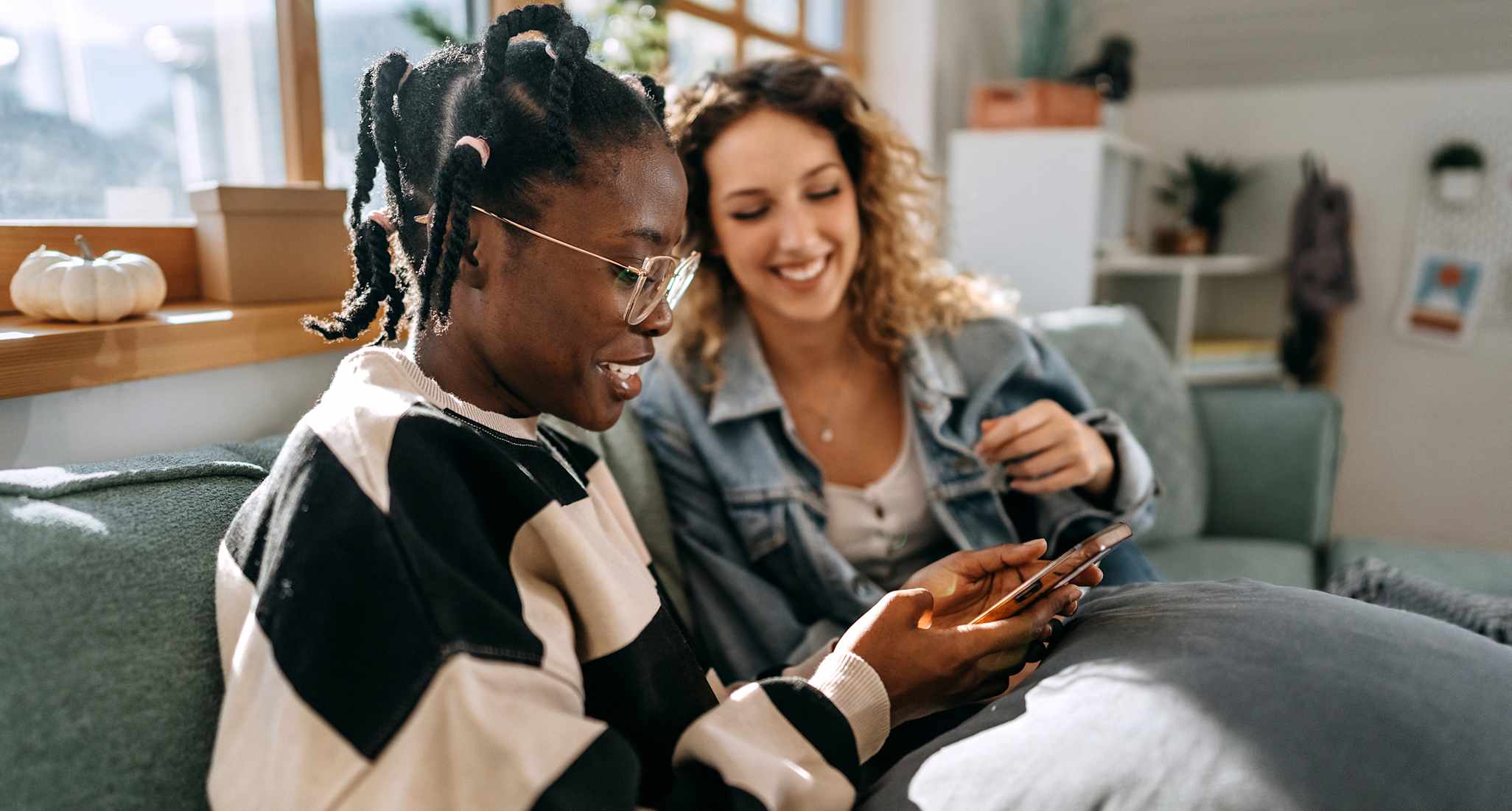 Zwei junge Frauen sitzen in ihrer gemeinsamen Wohnung auf dem Sofa und schauen zusammen aufs Handy.