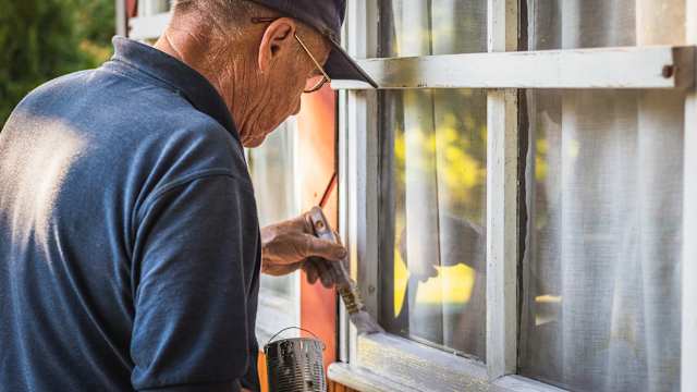 Un homme d’un certain âge repeint au pinceau le cadre extérieur d’une fenêtre en bois usée