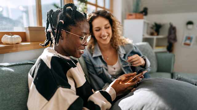 Zwei junge Frauen sitzen in ihrer gemeinsamen Wohnung auf dem Sofa und schauen zusammen aufs Handy.