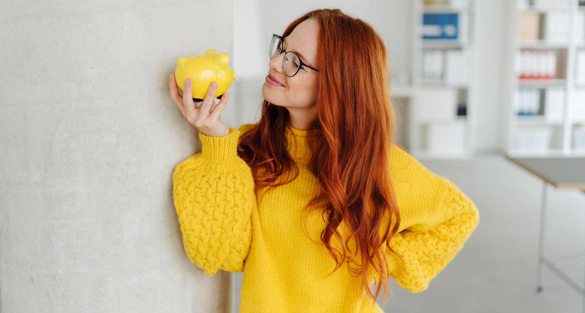 A woman in a yellow pullover is looking at a yellow piggy bank.