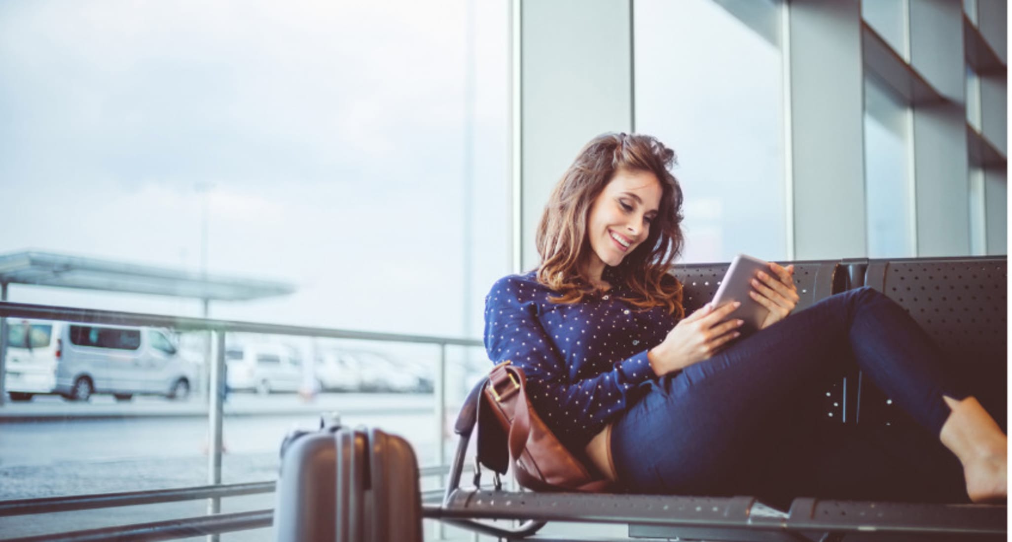 Eine Frau sitzt lächelnd am Flughafen und wartet auf ihren Flug. Sie liest ein E-Book, dass sie gratis heruntergeladen hat. 