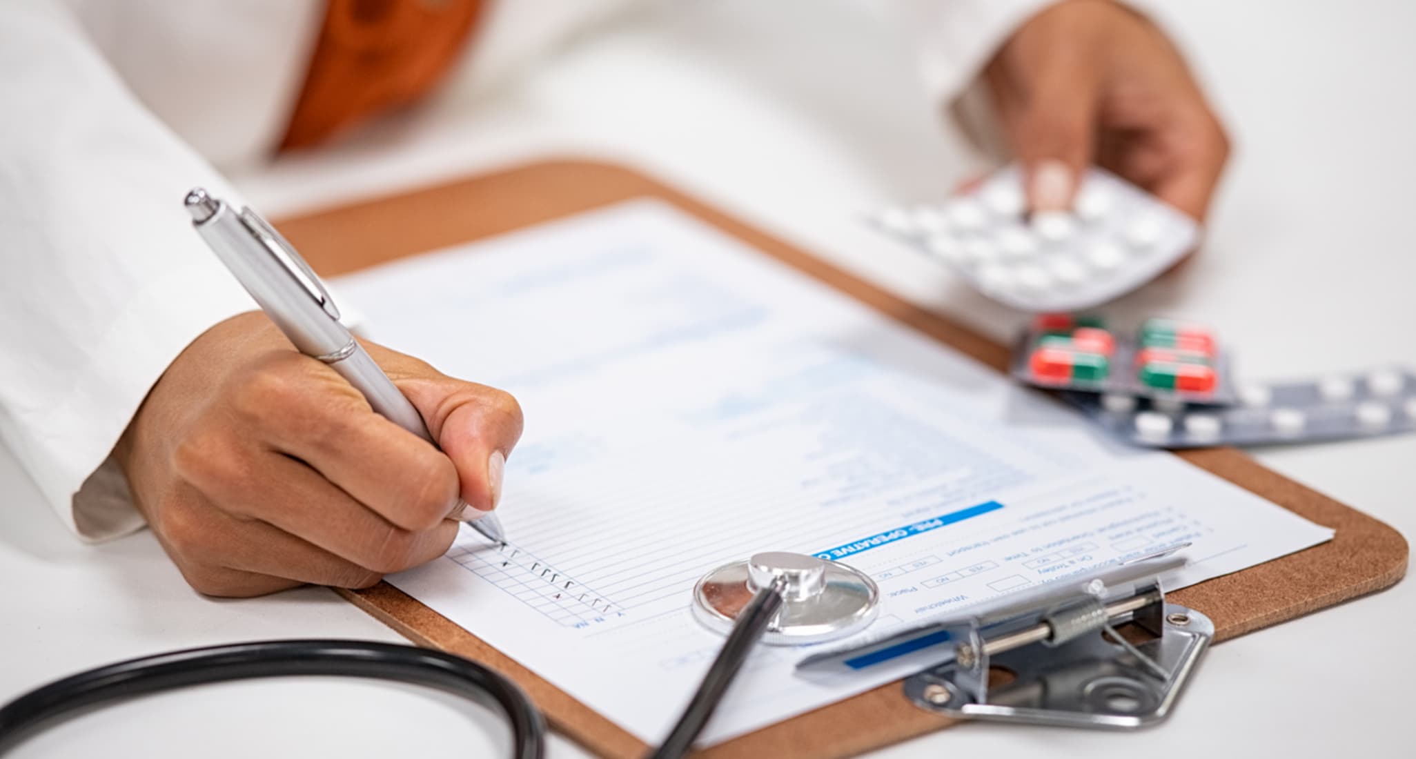 A woman writes on a sheet of paper on a clipboard. She has various pills in her left hand.