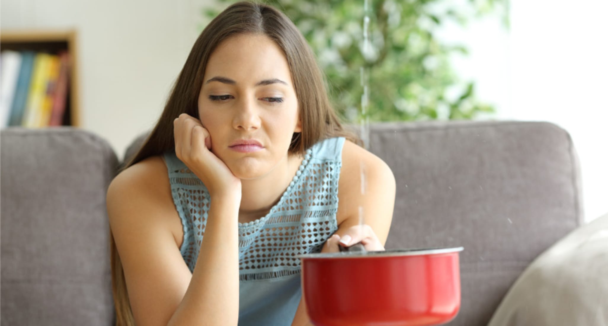 A young woman sits on a sofa. She is holding a pan in her left hand. It's catching water that is dripping from the ceiling. 