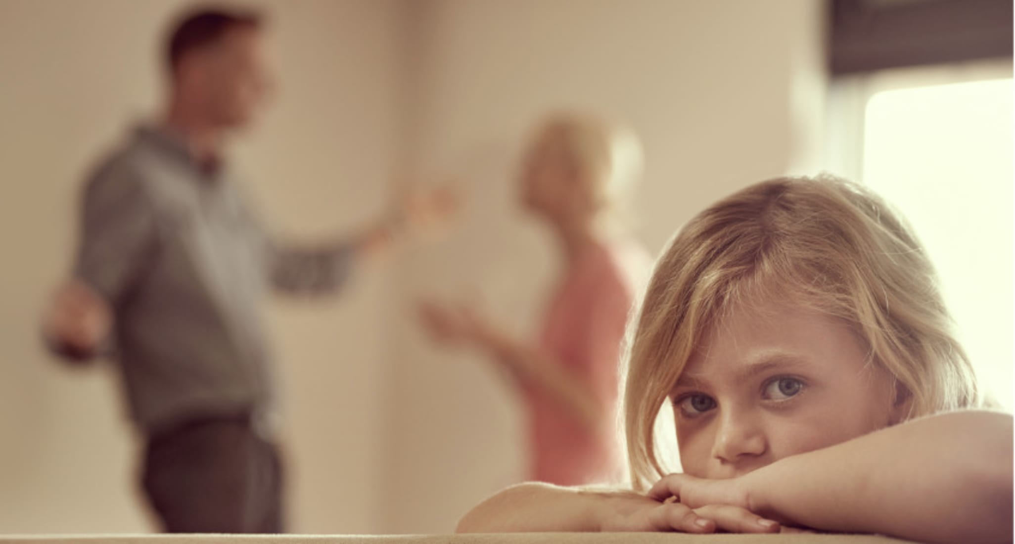 Una ragazza triste guarda nella telecamera. Sullo sfondo ci sono i suoi genitori che litigano.