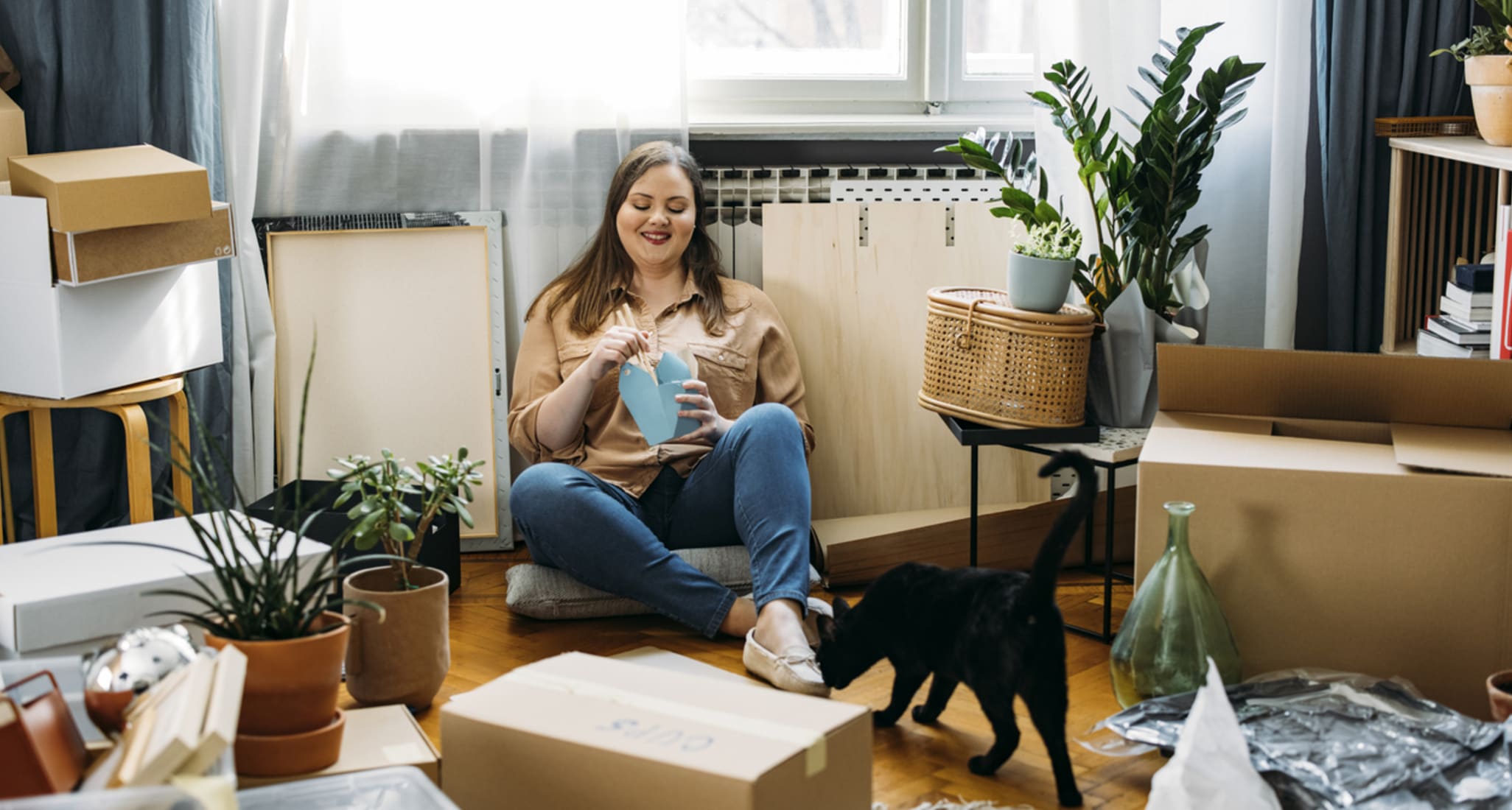 A young woman sits in her apartment after moving and eats food delivered to her home.