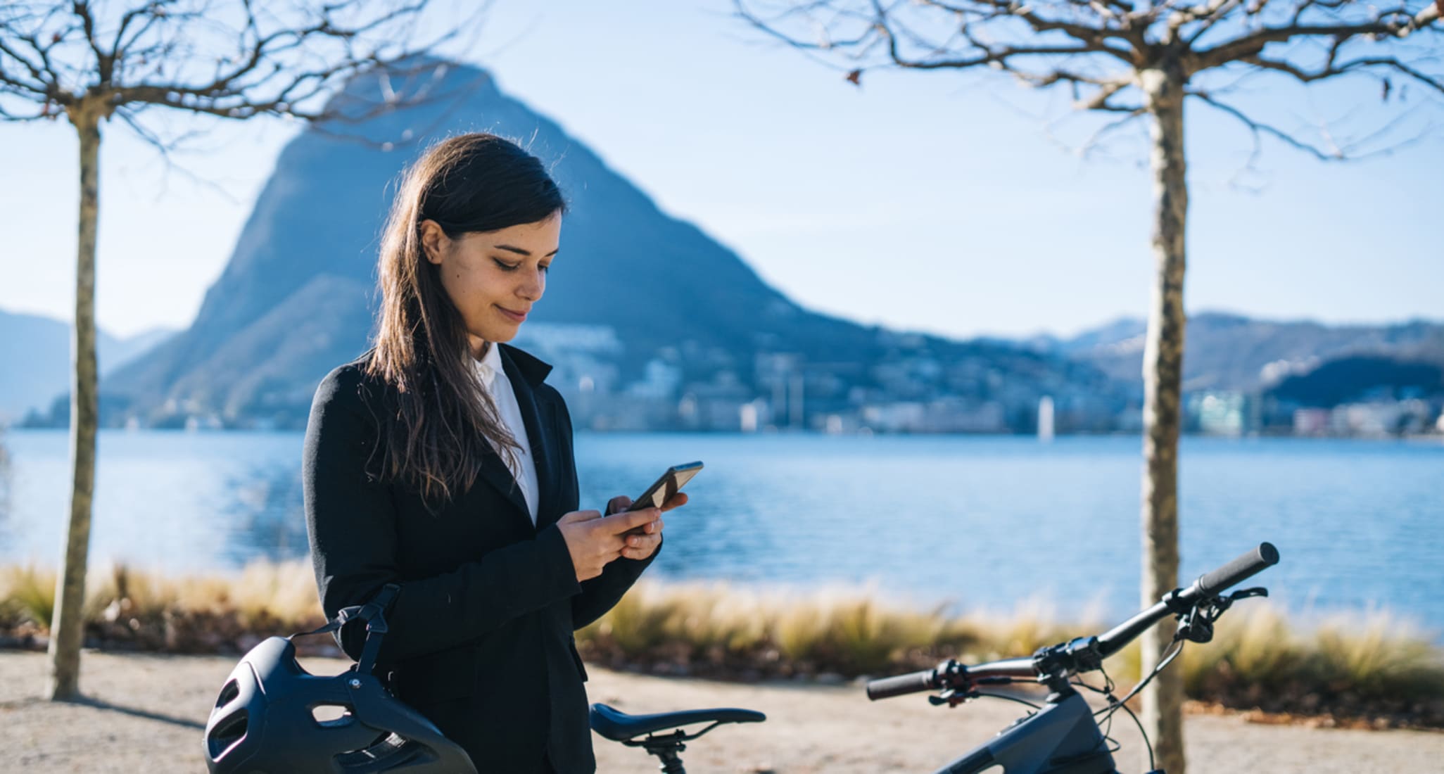 Eine Frau mit Smartphone in der Hand steht mit ihrem Fahrrad vor dem Luganersee.