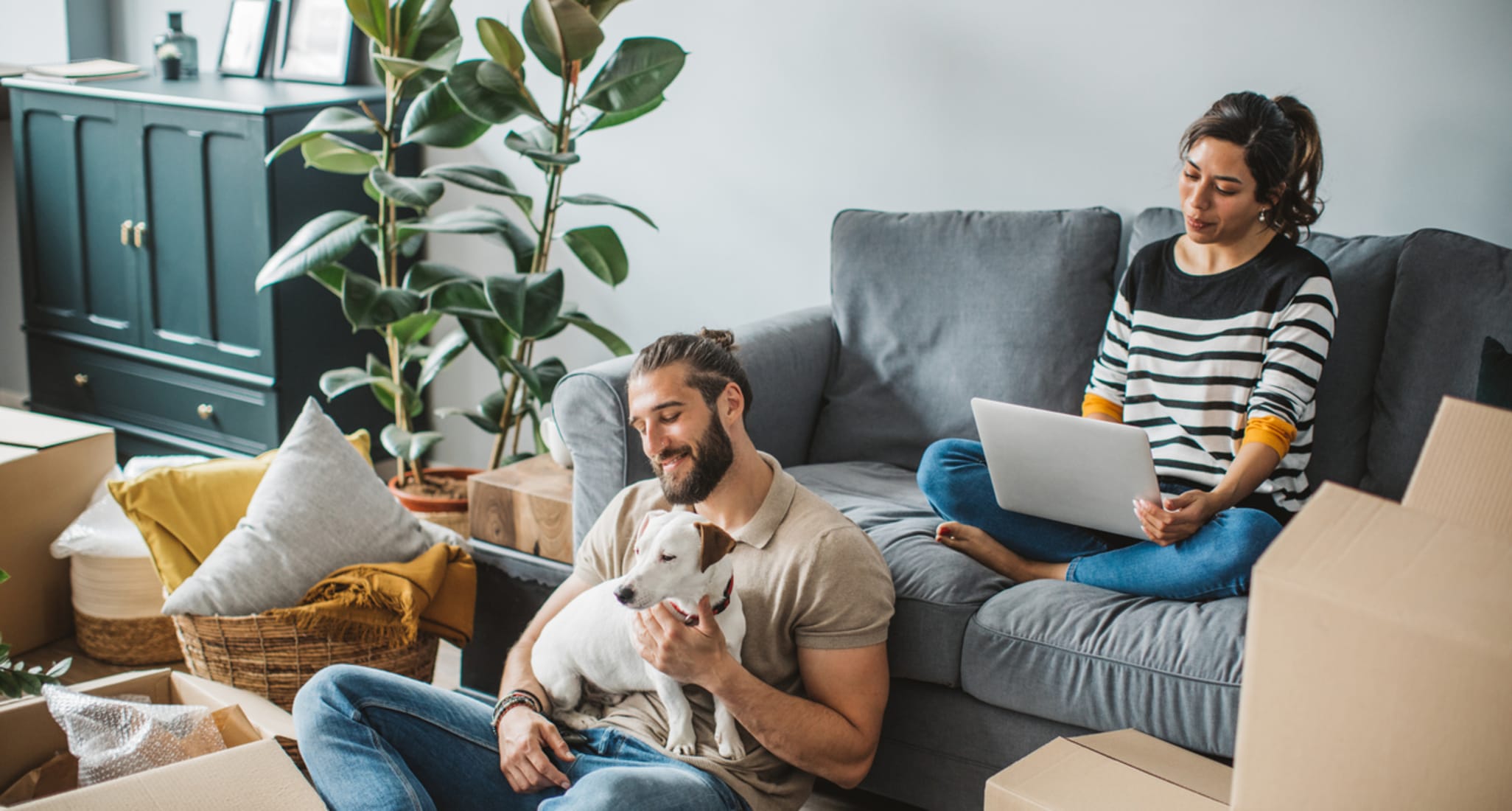 A couple sits between moving boxes. The woman sits on the sofa, the man on the floor with a dog on his lap.