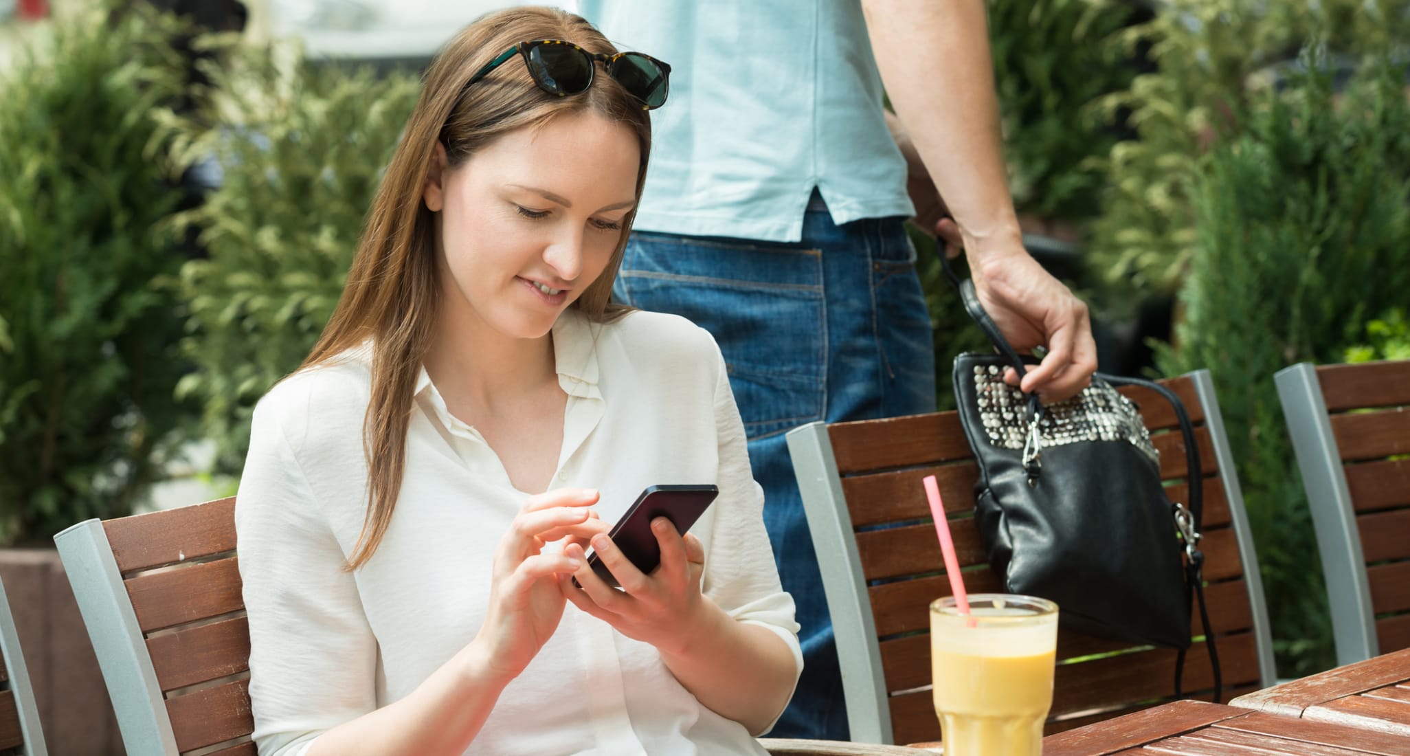 Una donna seduta a un tavolo in un caffè guarda il suo cellulare. Accanto a lei, qualcuno le ruba la borsa.