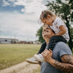 A man carries his daughter on his shoulders outside.