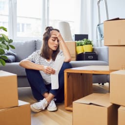 A worried young woman sits on the floor next to boxes. She is looking at a bill. 