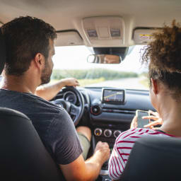 A couple sits in a car. The man looks at the street and the woman at her phone.