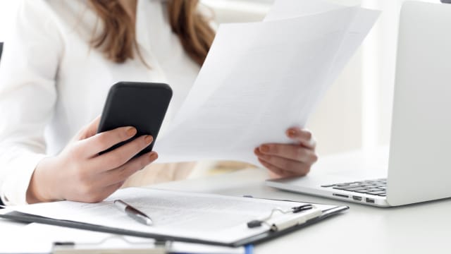 A woman at a computer holds her mobile phone and a contract in her hands. 